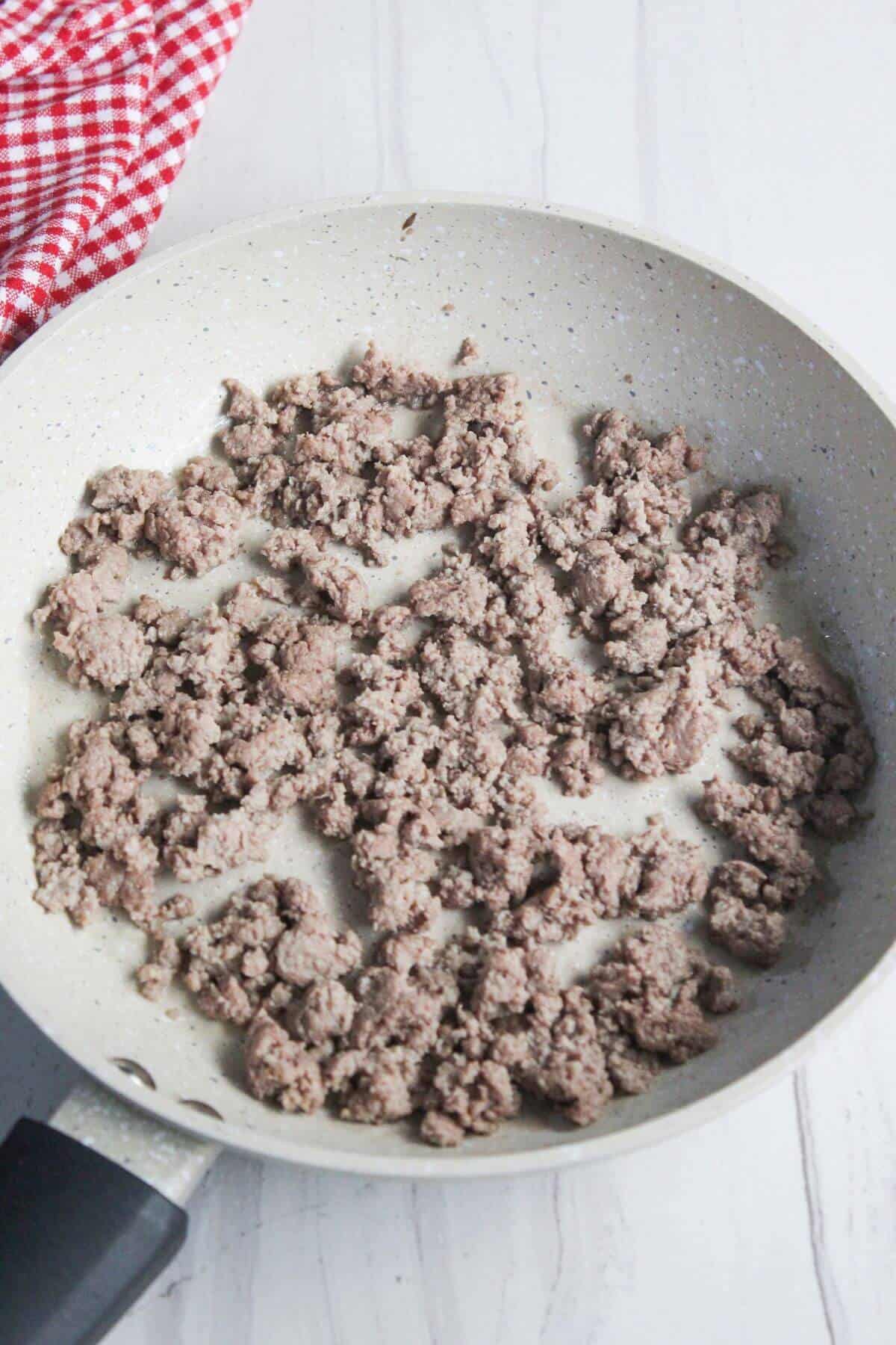 Ground meat cooking in a frying pan with a black handle on a white surface; red and white checkered cloth nearby.