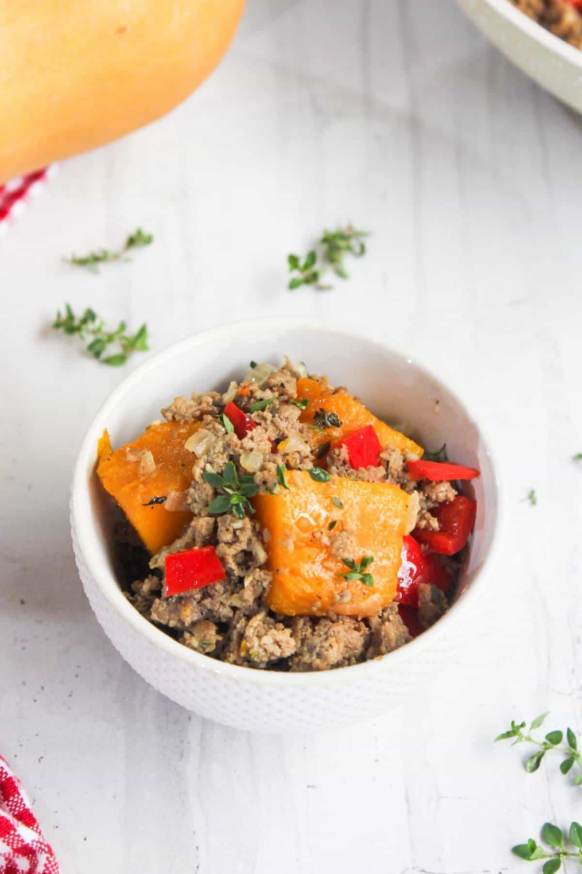 A white bowl containing cooked ground meat, pieces of butternut squash, red bell peppers, and garnished with fresh thyme, placed on a white surface.