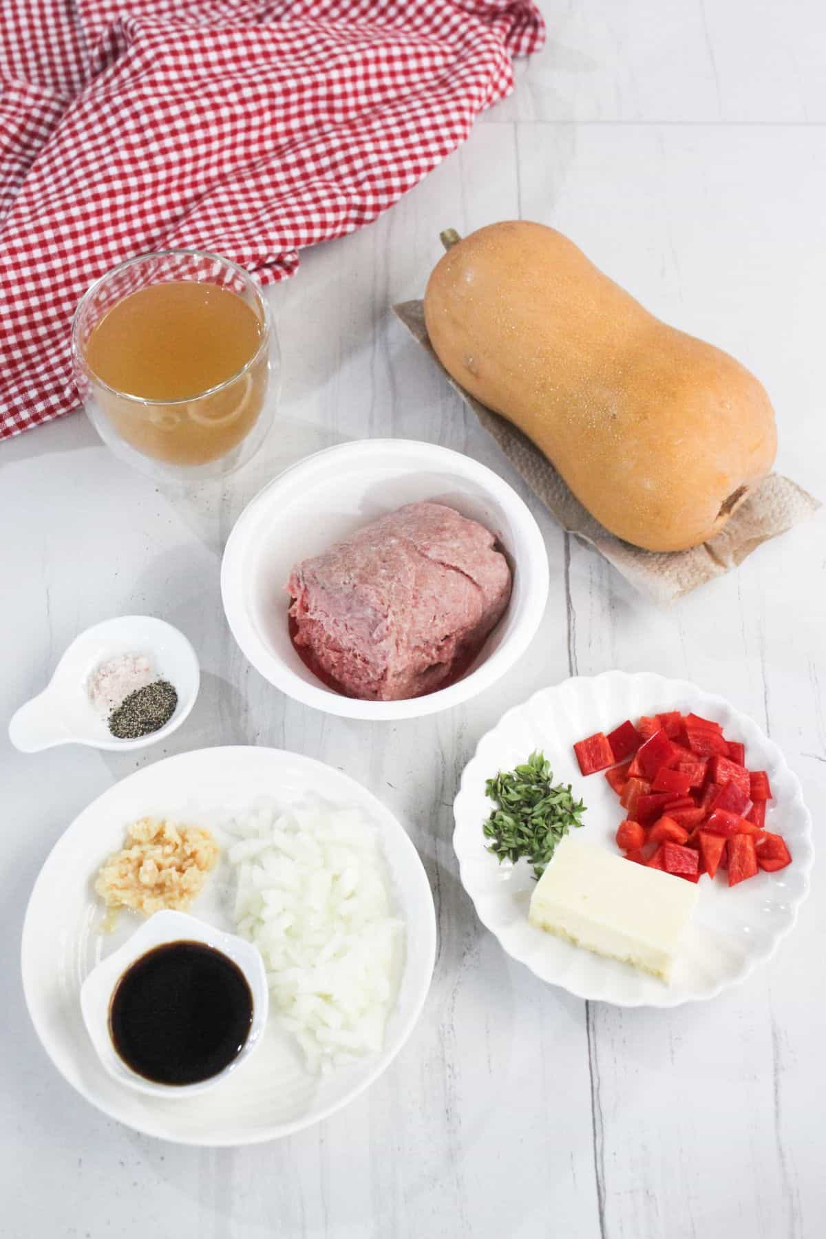 Ingredients on a table: ground meat, cubed red pepper, herbs, squash, glass of broth, chopped onion, soy sauce, minced garlic, butter, and a small dish of salt and pepper.