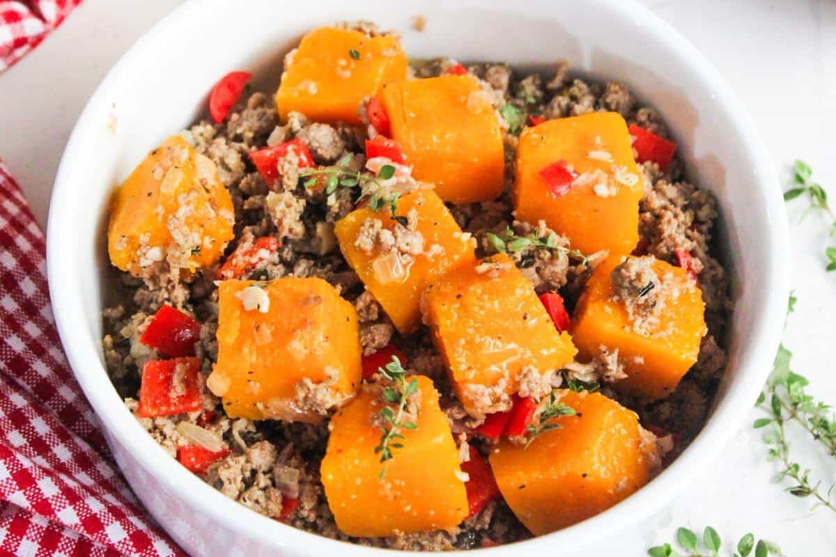 A bowl of cubed butternut squash, ground meat, and diced red peppers, garnished with herbs, next to a red checkered cloth.