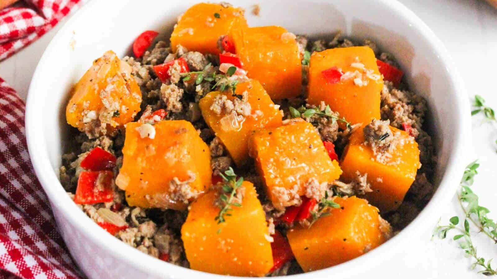 A bowl of cooked butternut squash and ground beef, garnished with herbs and diced red peppers.