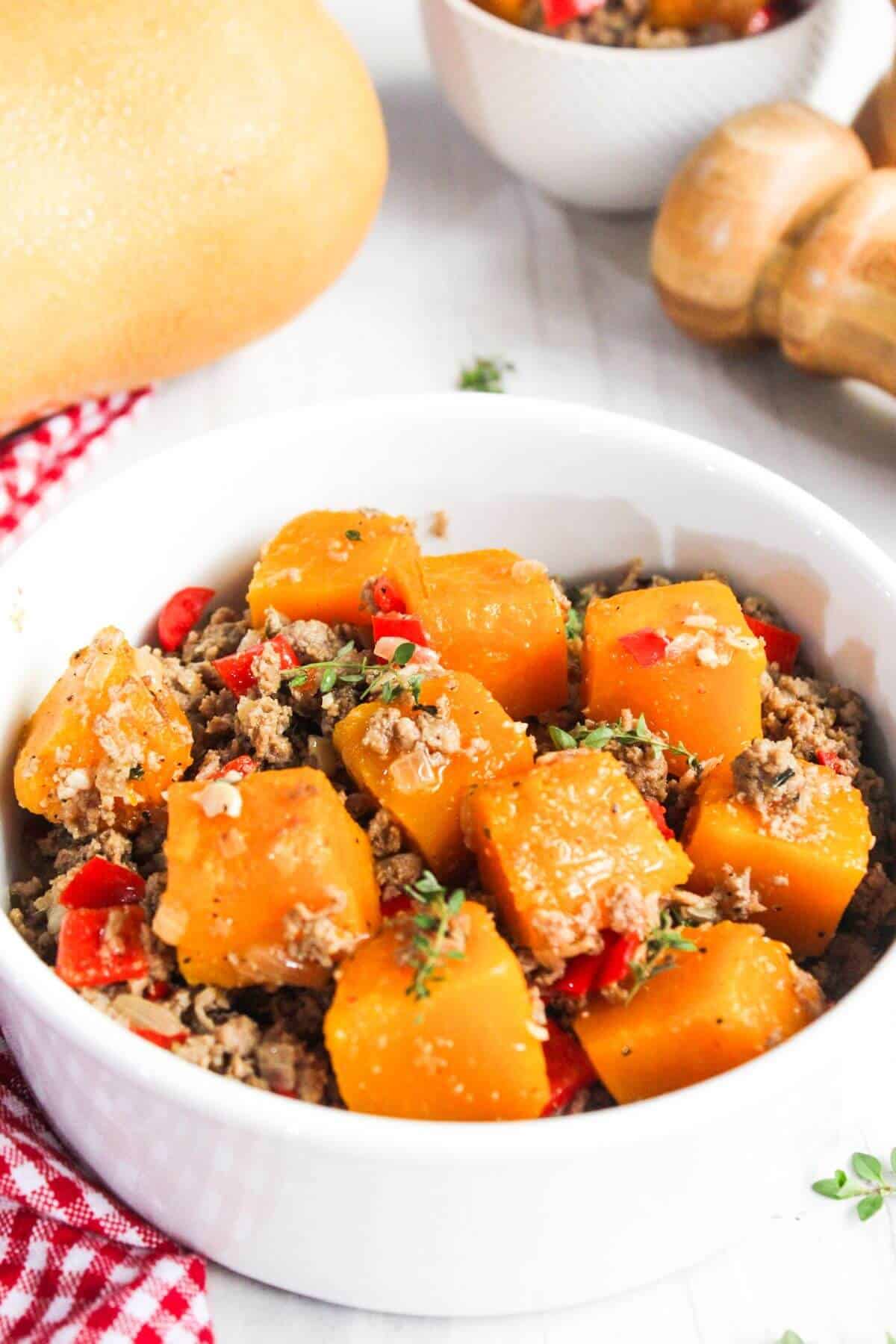 A white bowl filled with a dish of cubed orange squash, ground meat, red peppers, and herbs. A red checkered cloth, bread, mushrooms, and a small bowl in the background.