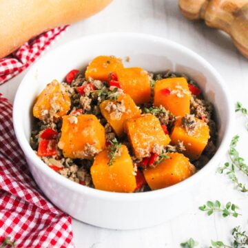 A white bowl filled with cooked pumpkin chunks, ground meat, and red peppers garnished with herbs, placed on a red-checked cloth.