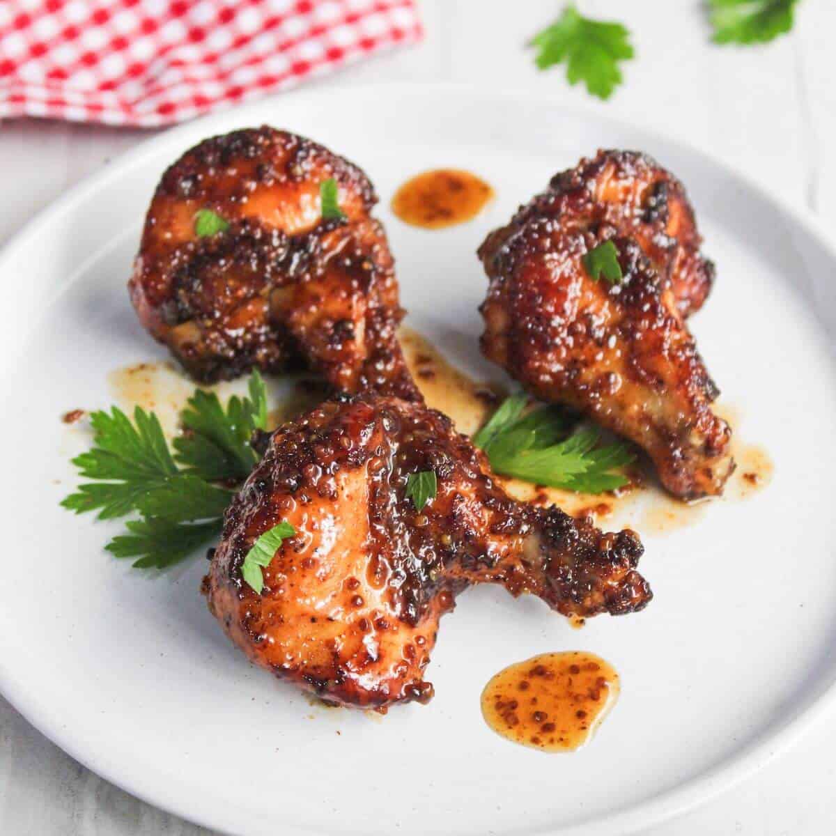 Three glazed chicken drumsticks garnished with green herbs on a white plate, alongside sauce droplets and parsley leaves.