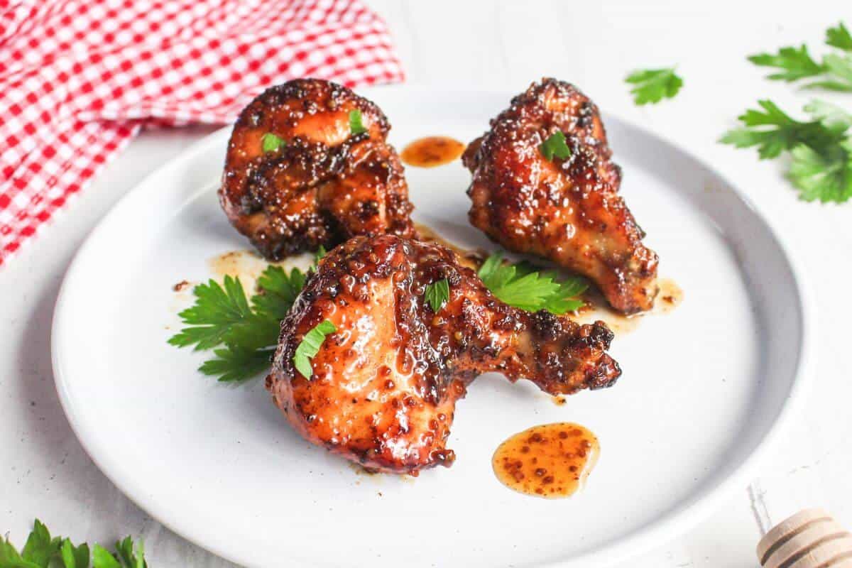 Three glazed chicken drumsticks on a white plate, garnished with parsley, placed next to a red checkered cloth.