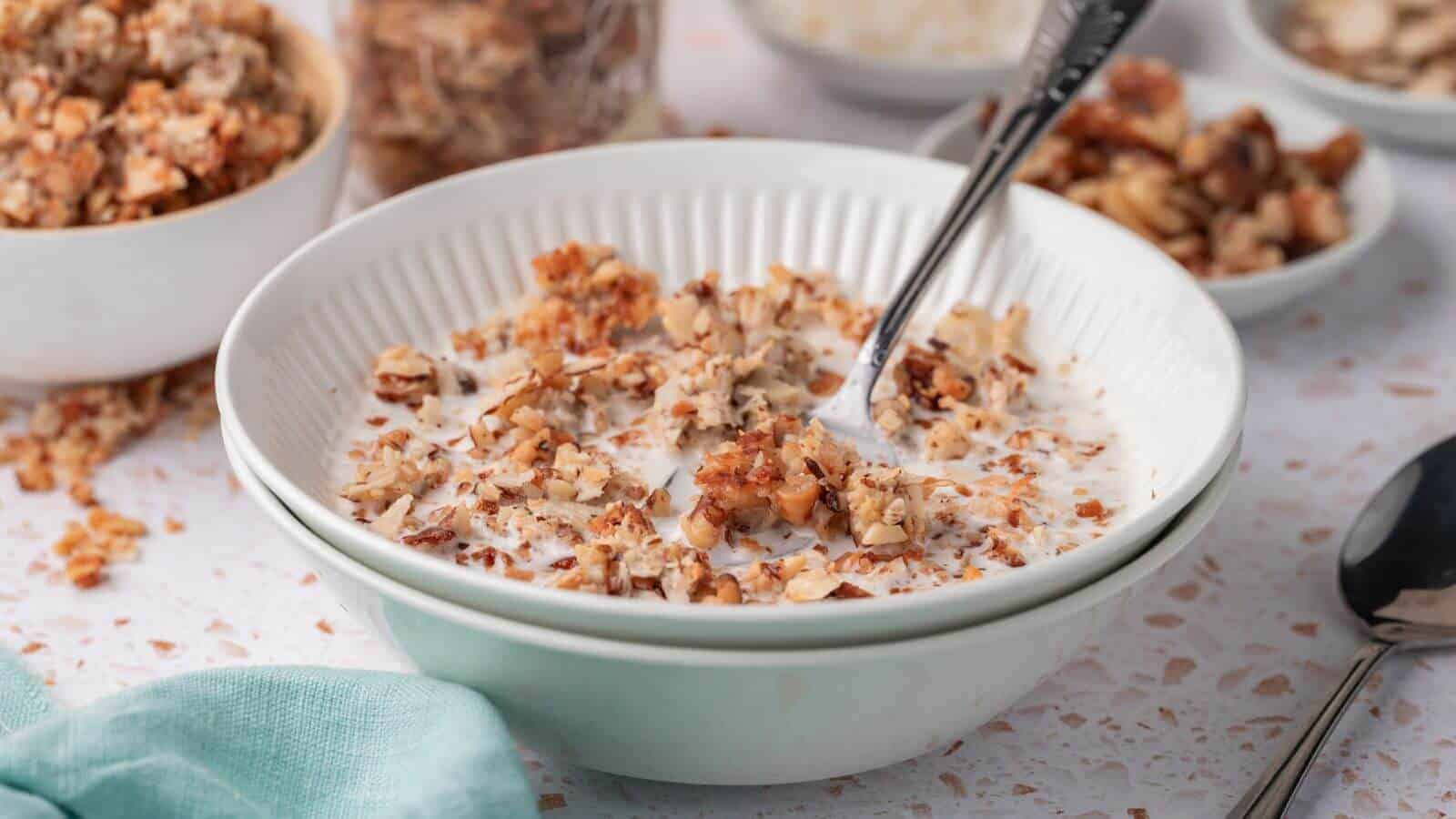 A bowl of cereal with milk and a spoon, surrounded by jars of granola on a light-colored tabletop.