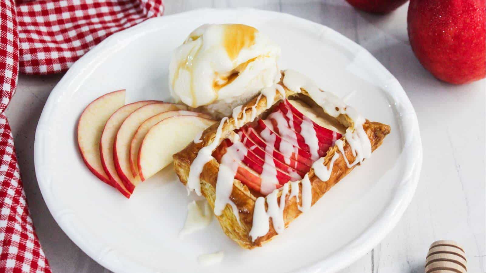 A dessert plate with apple slices, a pastry drizzled with icing, and a scoop of vanilla ice cream topped with syrup. A checkered cloth is nearby.