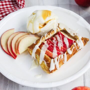Apple pastry with icing on a white plate, accompanied by sliced apples and a scoop of ice cream topped with syrup.
