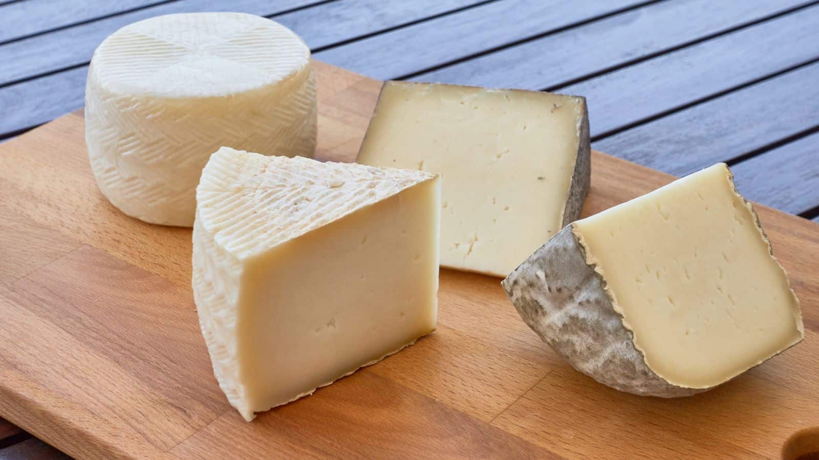A wooden board with four various cheese wedges, differing in texture and rind color, placed on a table.