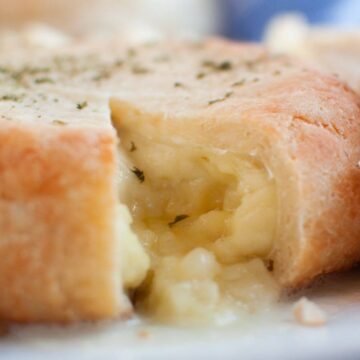 A baked wheel of Brie cheese wrapped in pastry partially cut open, revealing melted cheese inside. Surrounded by pieces of bread and crackers.