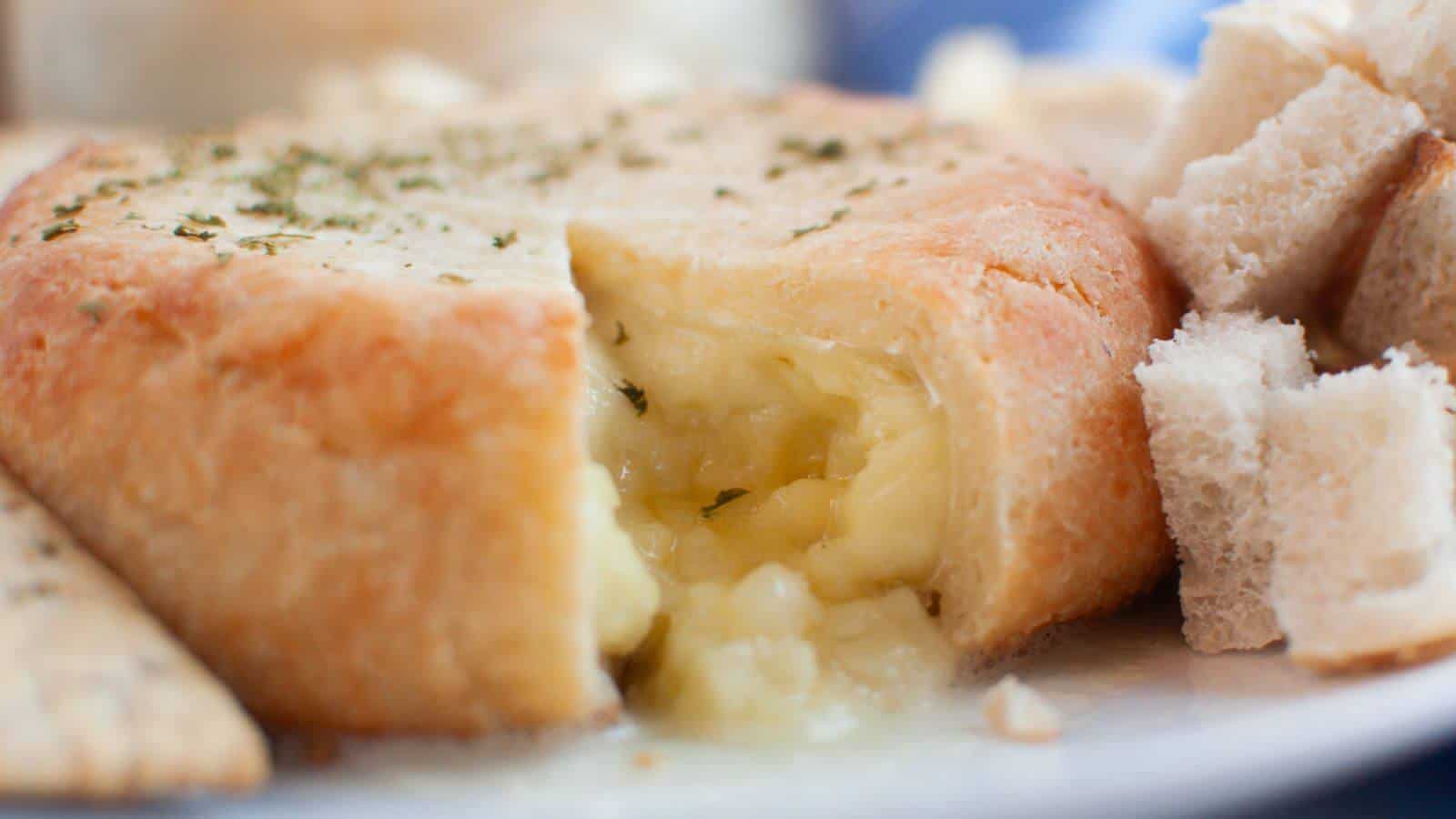 A baked wheel of Brie cheese partially cut open, revealing melted cheese inside. Surrounded by pieces of bread and crackers.
