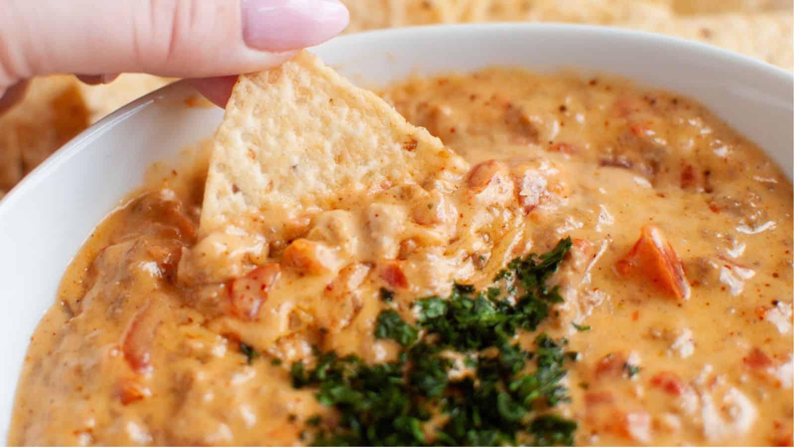 A hand dipping a tortilla chip into a bowl of cheesy dip with diced tomatoes and herbs.