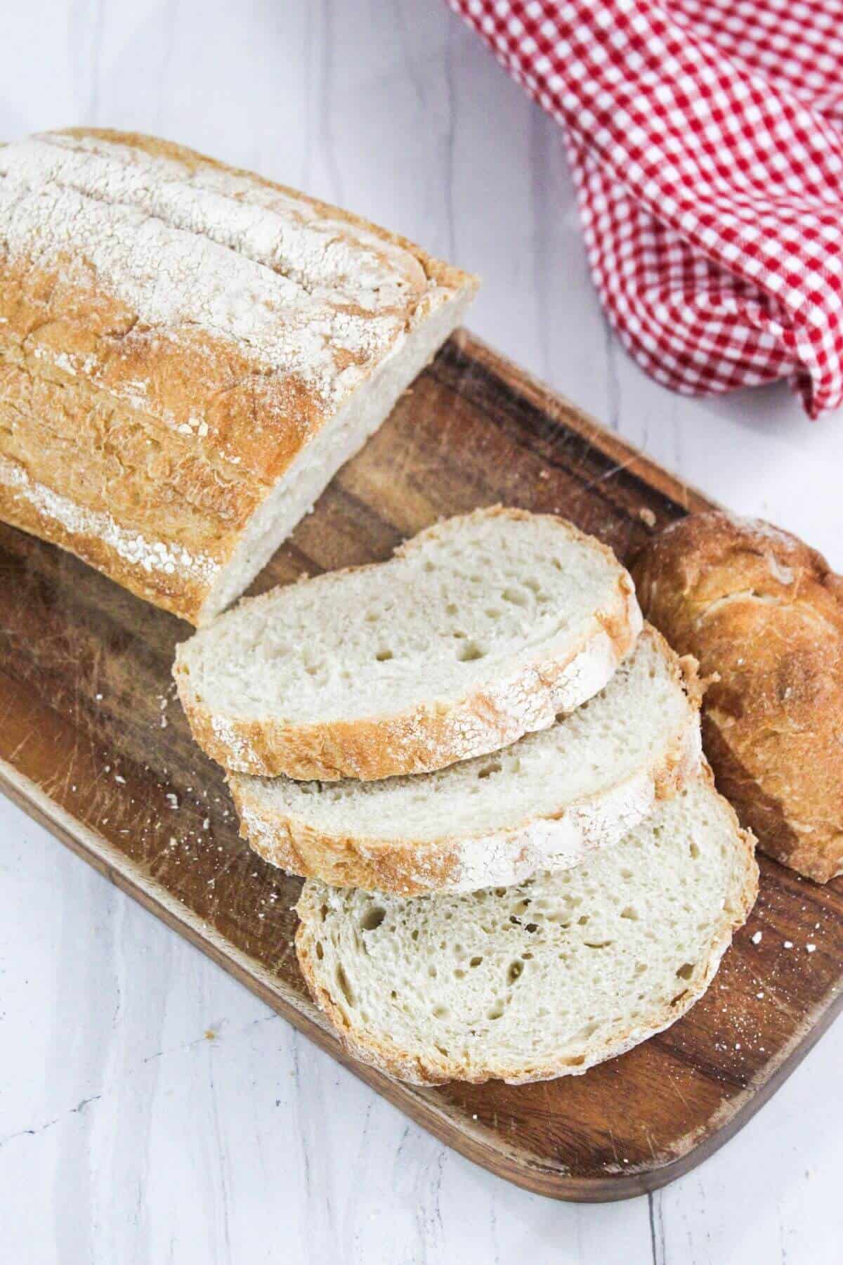 Sliced loaf of white bread on a wooden board with a red checkered cloth in the background.