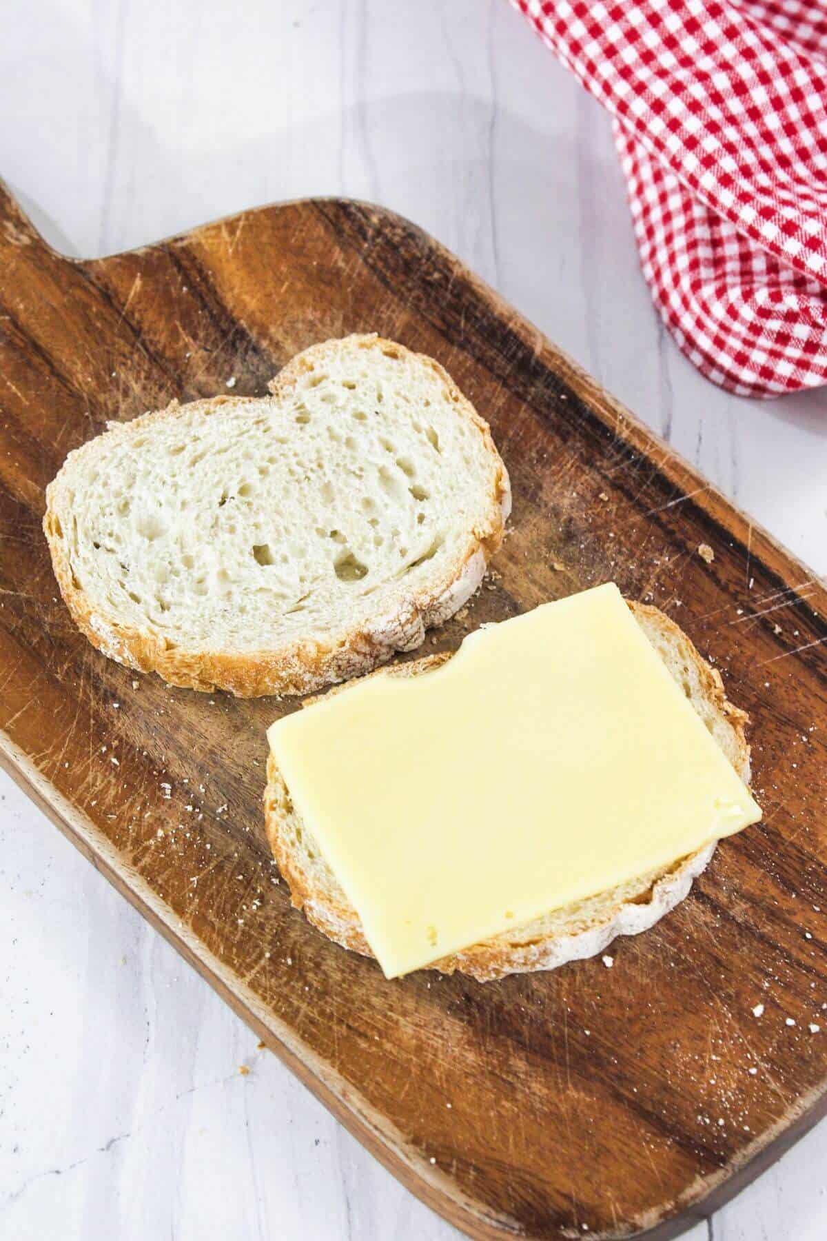 Two slices of bread on a wooden board, one topped with a slice of cheese. A red and white checkered cloth is partially visible.
