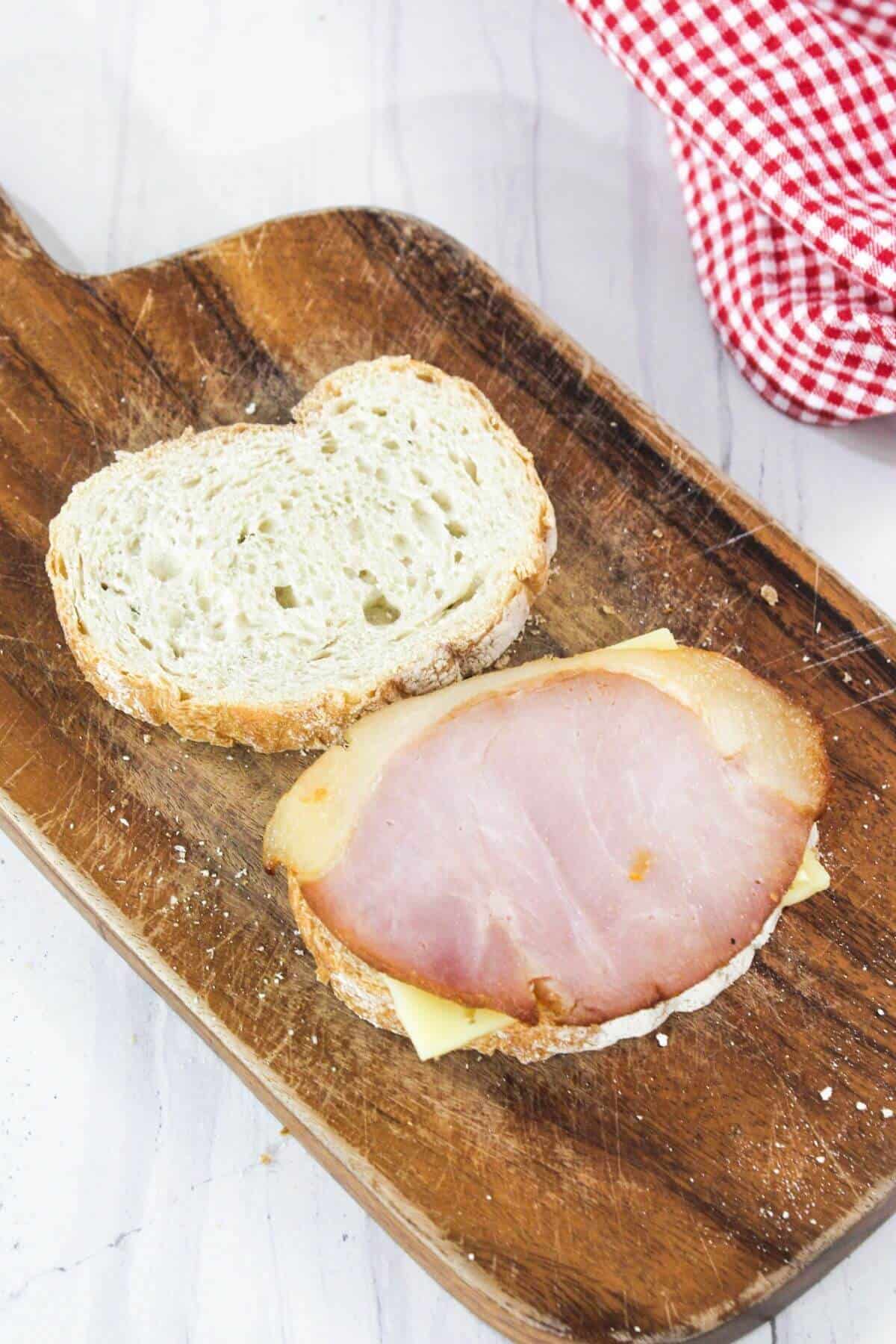 Slice of bread with cheese and ham on a wooden board, accompanied by a second slice of bread partially covering the ham. A red and white checkered cloth is nearby.