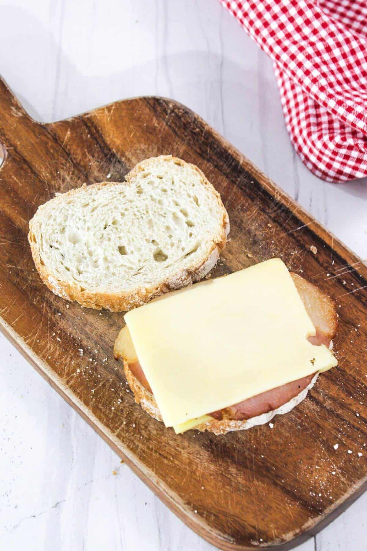 A sandwich in progress on a wooden board, featuring a slice of cheese on top of ham and bread, with a red checkered cloth nearby.