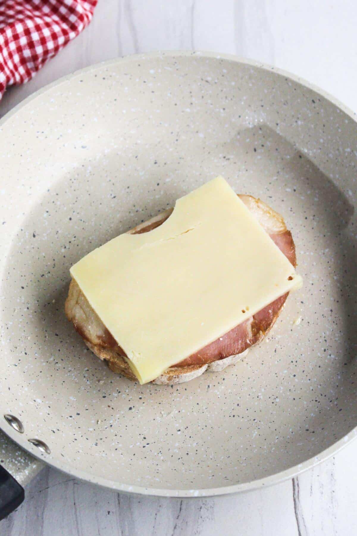 Slice of cheese on a piece of ham and bread in a frying pan with a red check towel nearby.