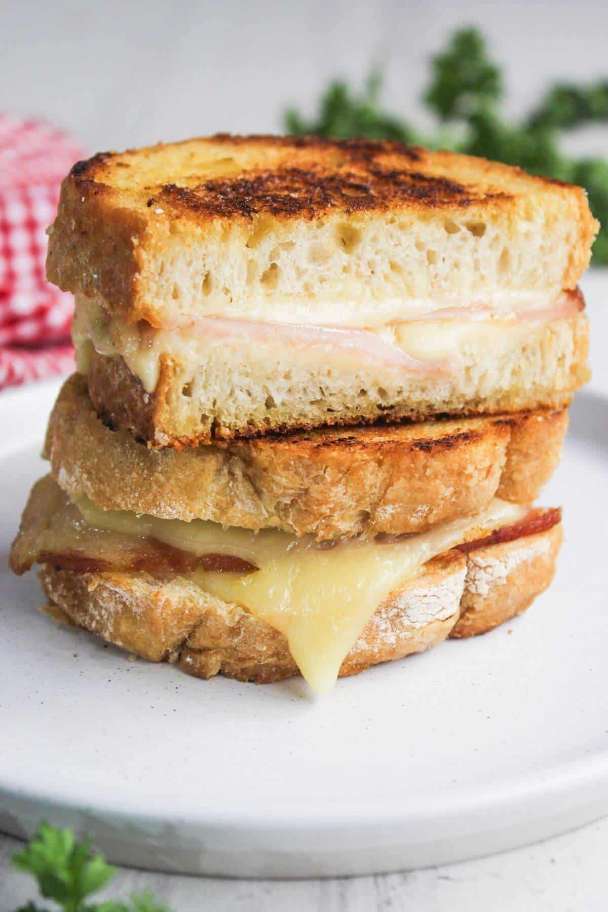 A grilled cheese sandwich stacked in halves on a white plate, with melted cheese oozing out. A red and white checkered cloth is in the background.