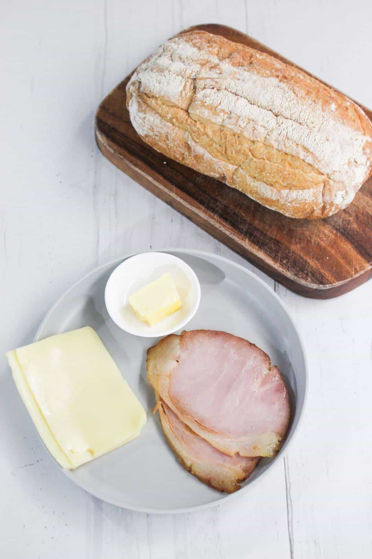 Plate with sliced ham, cheese, and butter next to a wooden board with a loaf of bread.