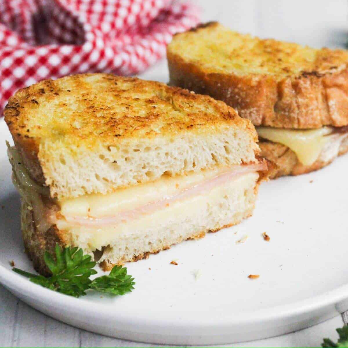 Grilled cheese and ham sandwich cut in half on a white plate, with a sprig of parsley and a red checkered cloth in the background.