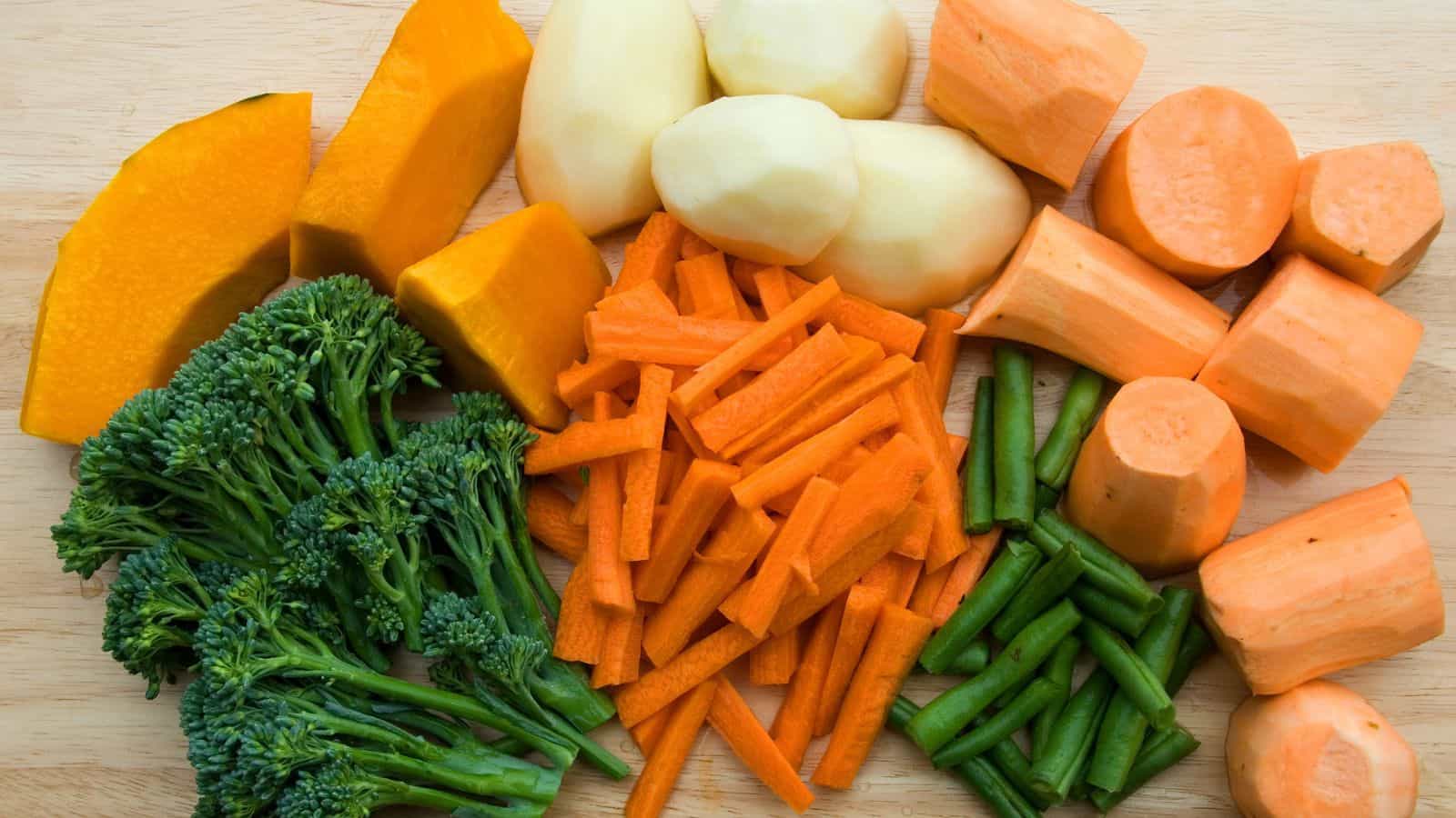 Variety of chopped vegetables on a wooden board: pumpkin wedges, peeled potatoes, sweet potato pieces, broccoli florets, carrot sticks, and green bean segments.