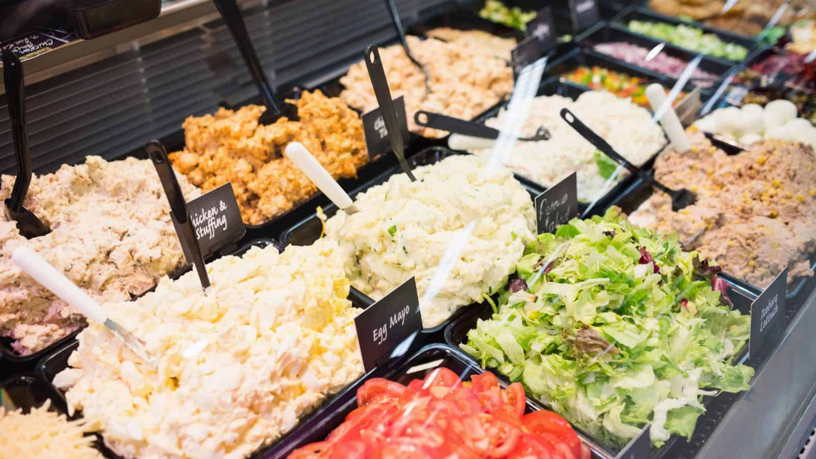 Display of various deli salads, including egg salad, chicken salad, pasta salad, and mixed greens, in black trays with serving spoons and labels.