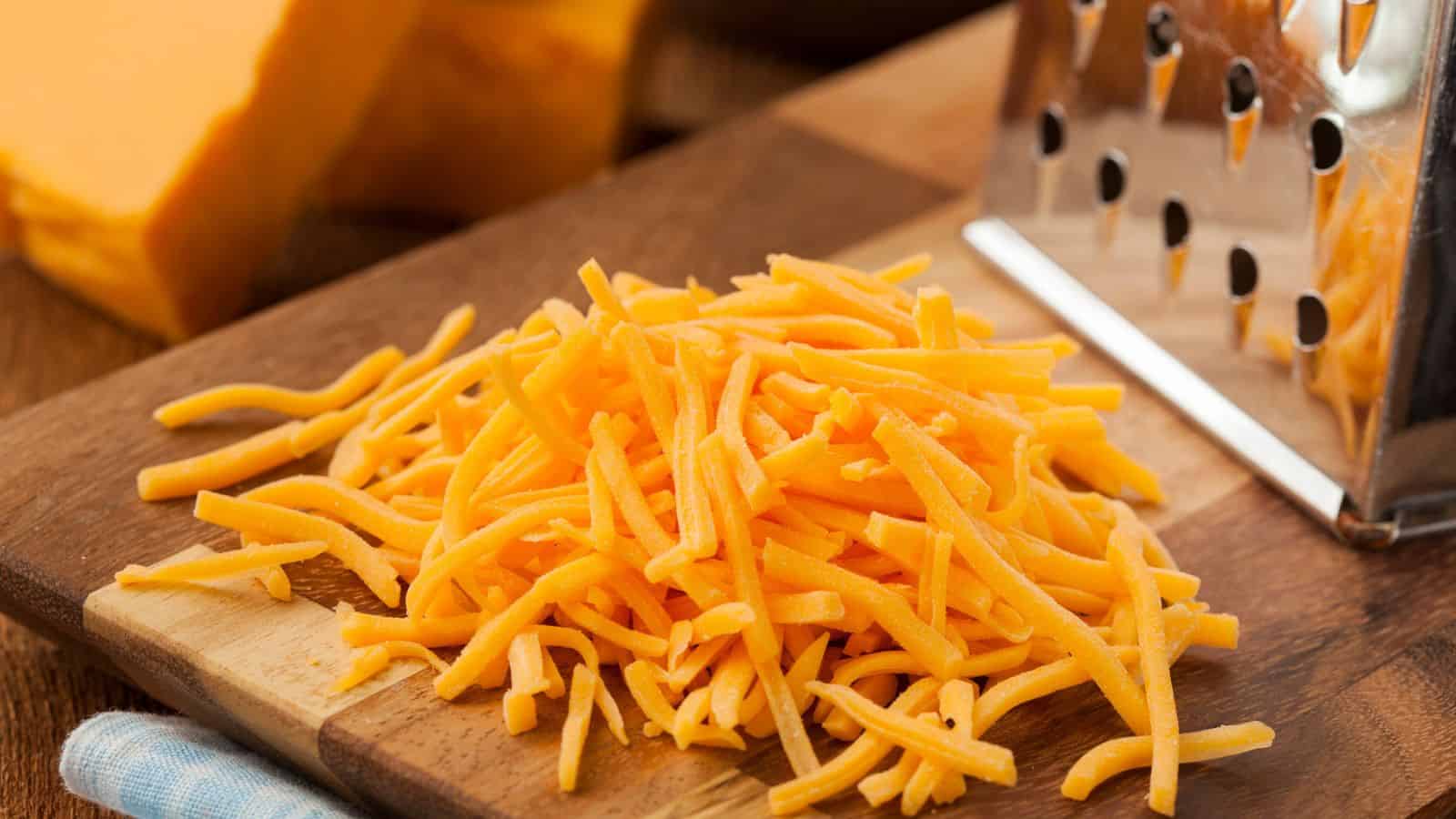 Shredded cheddar cheese on a wooden cutting board next to a metal grater.