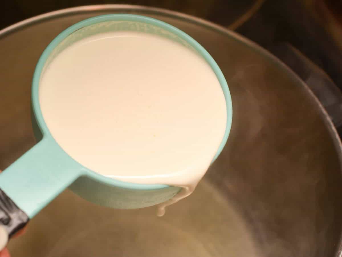 A light blue cup pouring milk into a stainless steel pot on a stove.