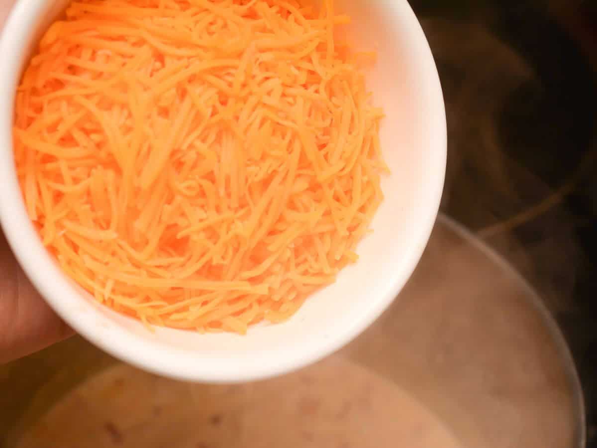 A hand holds a white bowl containing shredded orange cheese over a pot of soup.