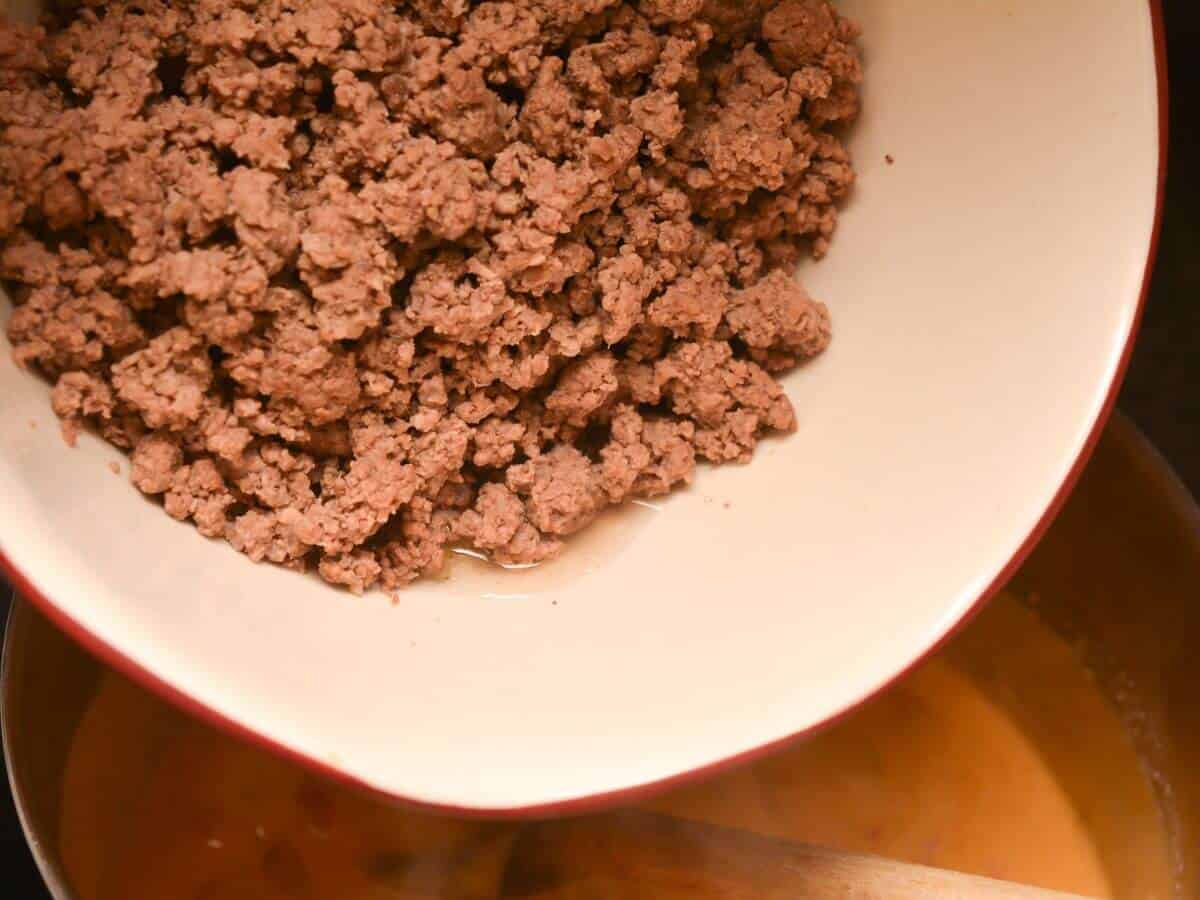 Ground beef being added from a white bowl into a pot with a wooden spoon stirring a liquid mixture.
