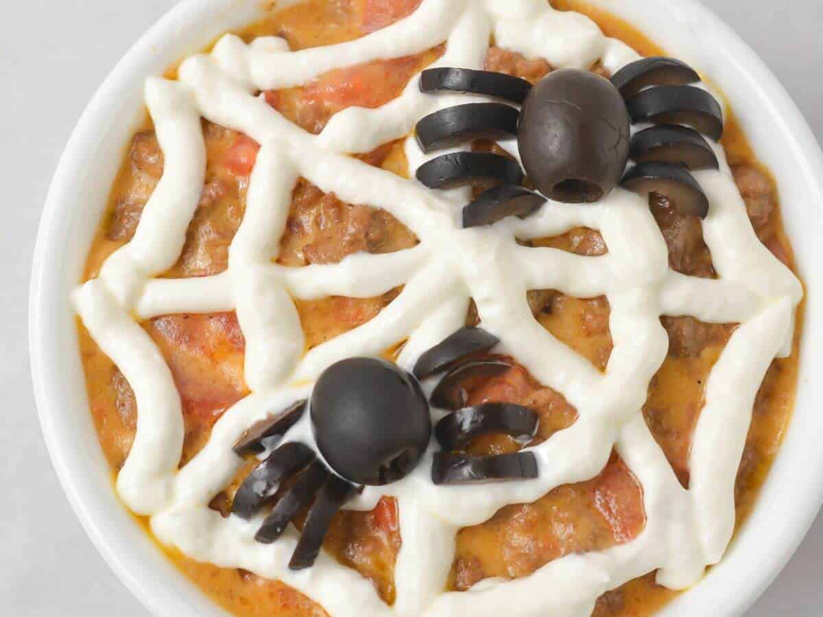 A bowl of dip decorated with sour cream in a spiderweb pattern and topped with two olive spiders.