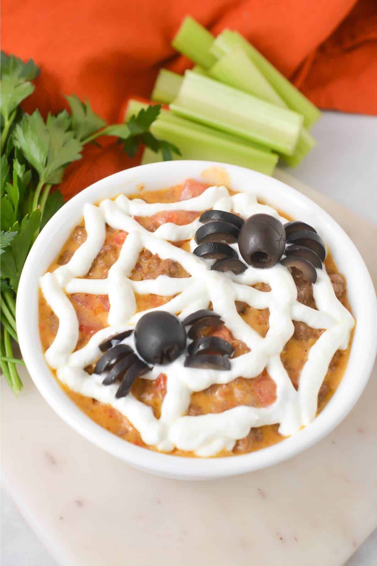 A bowl of dip topped with sour cream and olive spiders, garnished with celery and parsley, on a white surface with an orange cloth in the background.