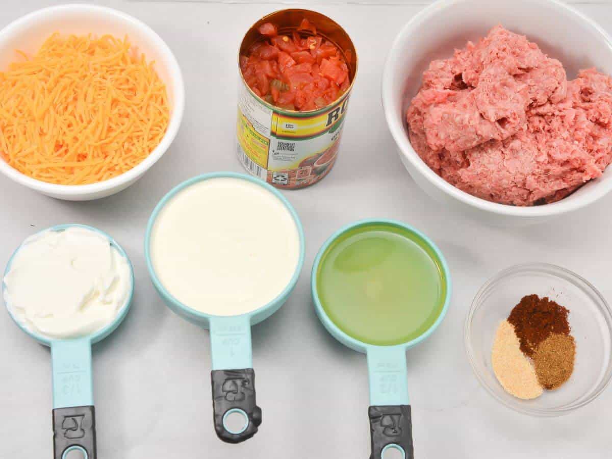 Ingredients for a dip: shredded cheese, canned tomatoes, ground beef, sour cream, green chili sauce, and seasonings in bowls and measuring cups on a white surface.