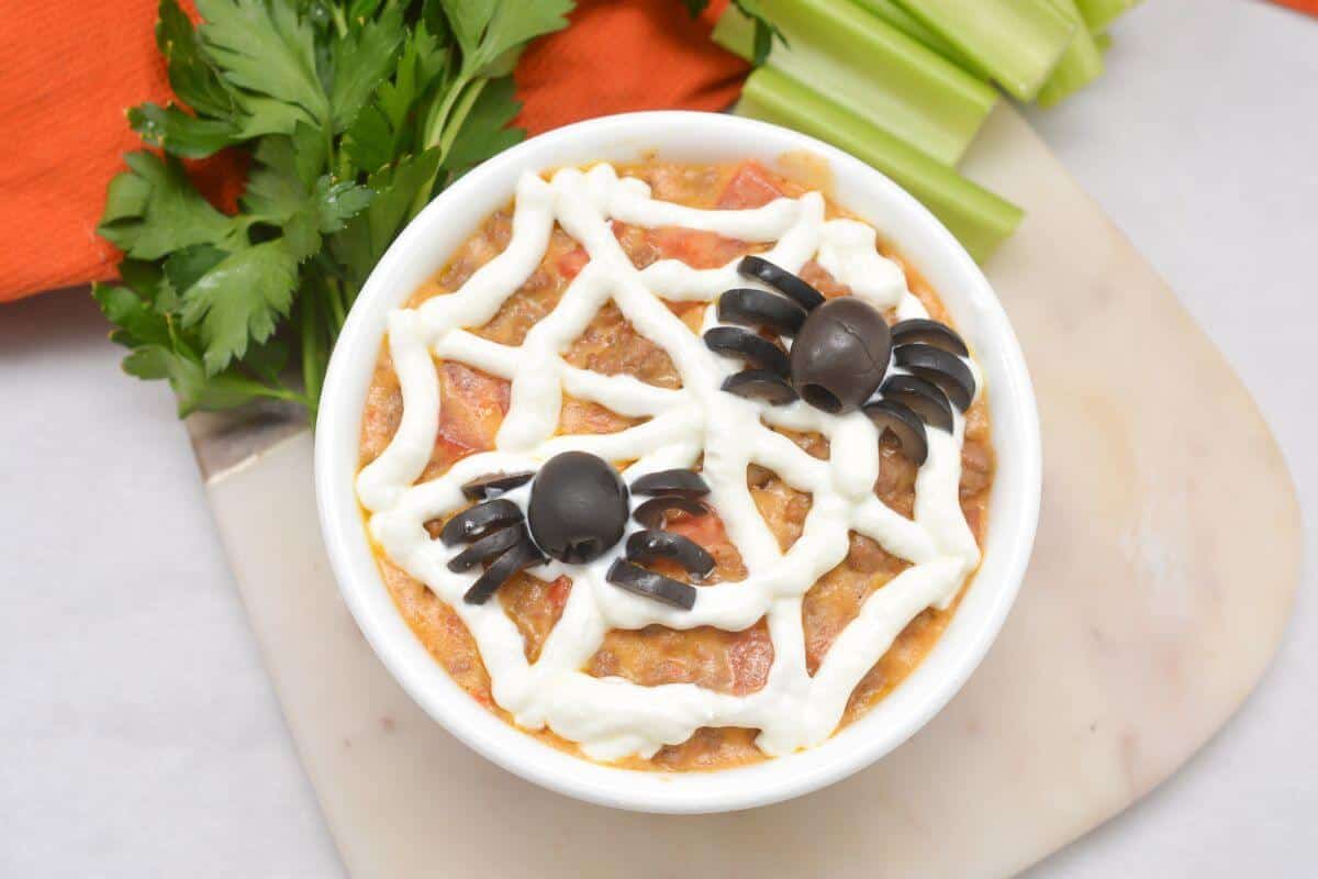 A bowl of dip topped with a spider web design made of sour cream and olive spiders, placed on a marble board with parsley, celery, and an orange napkin nearby.