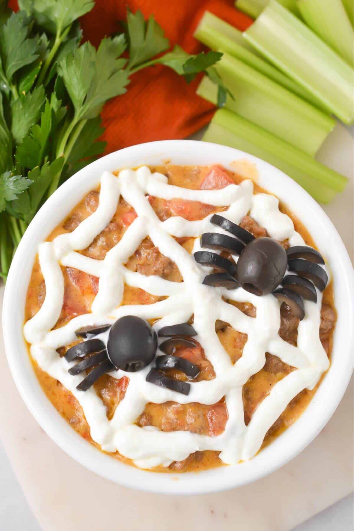 A bowl of dip topped with olives styled as spiders and a web design made of sour cream, placed next to parsley and celery sticks.
