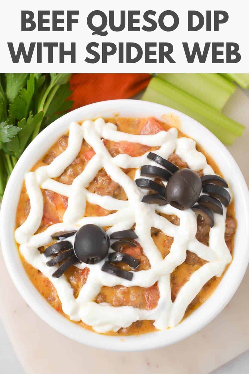 A bowl of beef queso dip topped with a sour cream spider web design and two olive spiders. Celery, carrots, and parsley are in the background.