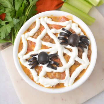 A bowl of dip with spider-shaped olives and sour cream web decoration, garnished with parsley and celery sticks on the side.
