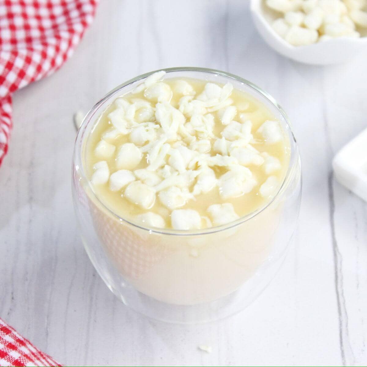 A glass of atole topped with small marshmallows on a white wooden table, next to a red checkered cloth and a small dish containing more marshmallows in the background.