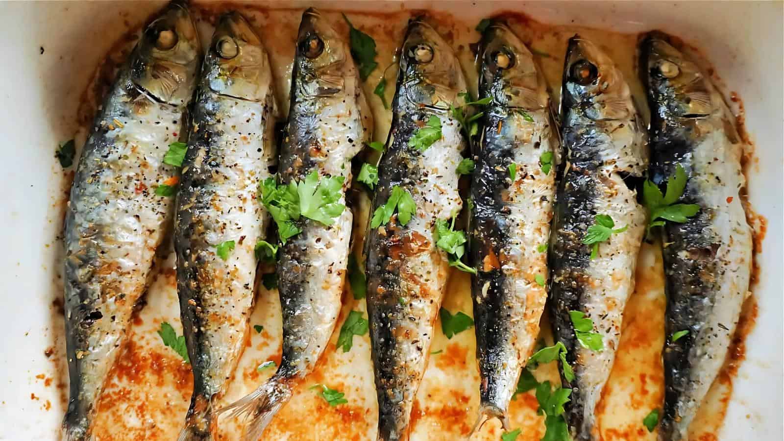 Eight seasoned and cooked fish laid out in a baking dish, garnished with herbs and black pepper.