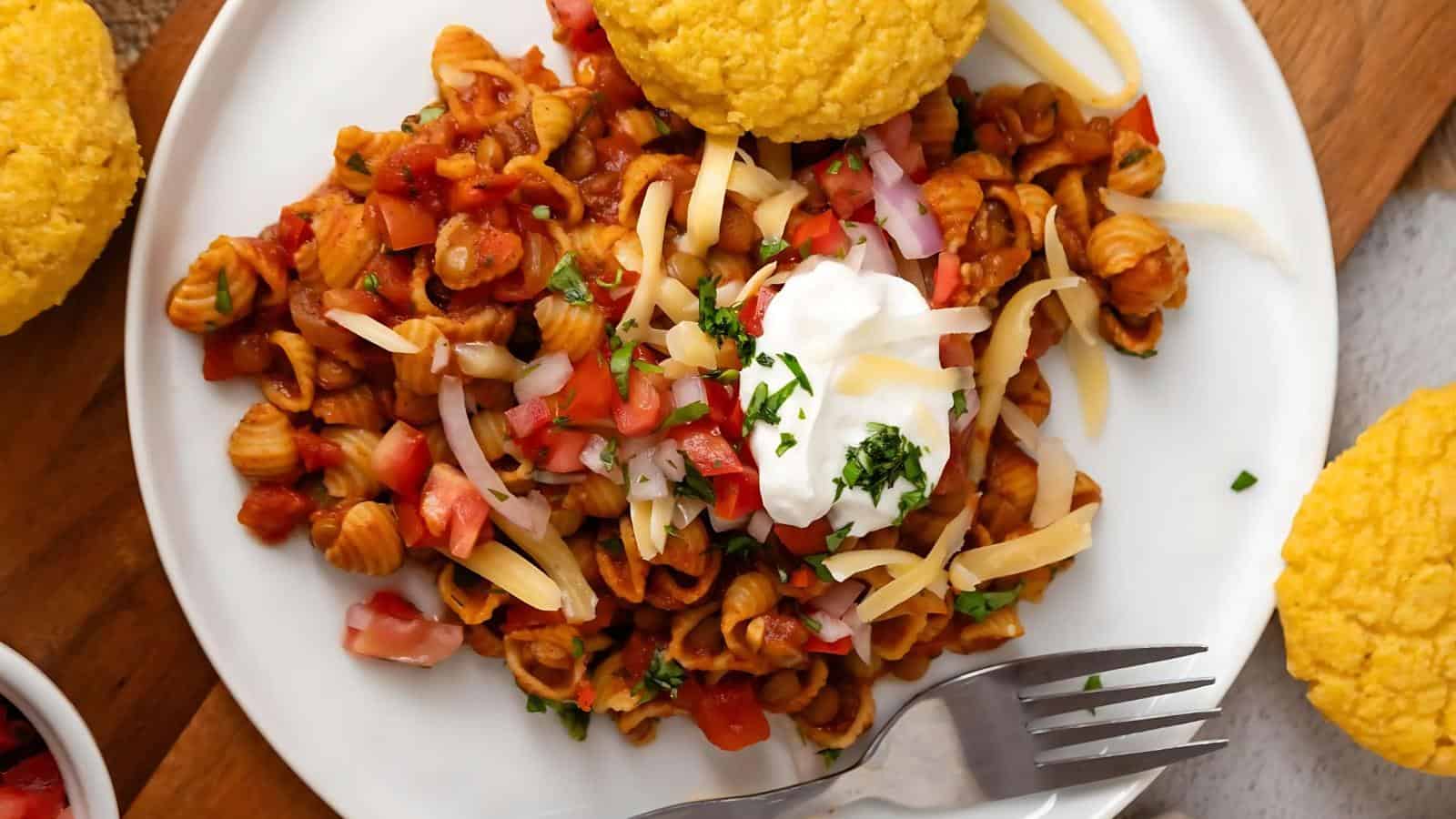 A plate with pasta topped with diced tomatoes, grated cheese, sour cream, and herbs, accompanied by two corn muffins on the side. A fork is placed on the plate.