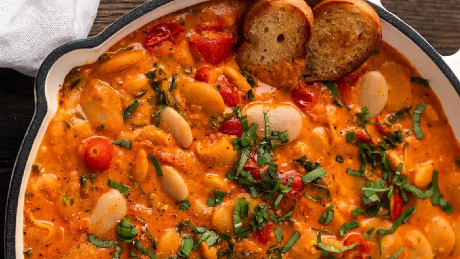 Close-up of a pan containing creamy tomato and bean stew, garnished with chopped herbs. Two toasted bread slices rest on the edge.