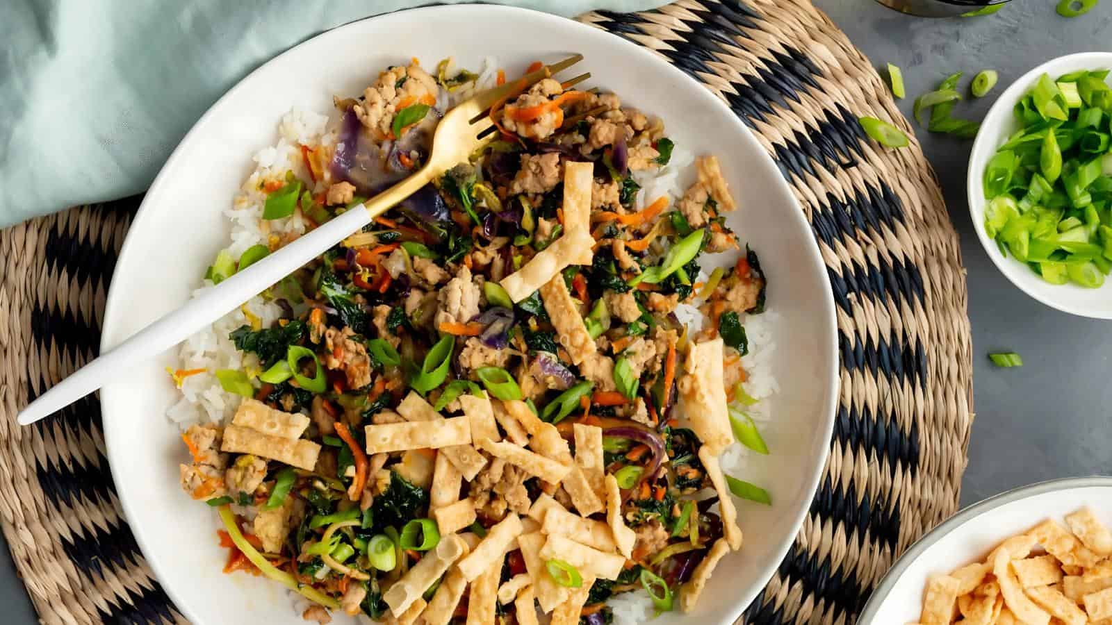 A plate of stir-fried vegetables and chicken over rice, garnished with green onions and crispy wonton strips, with a fork on the side.