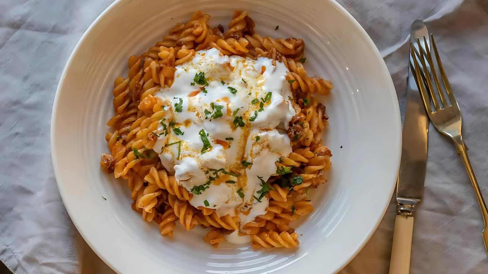 A plate of fusilli pasta with a creamy sauce and herbs is served on a white plate. A fork and knife are placed beside the plate.