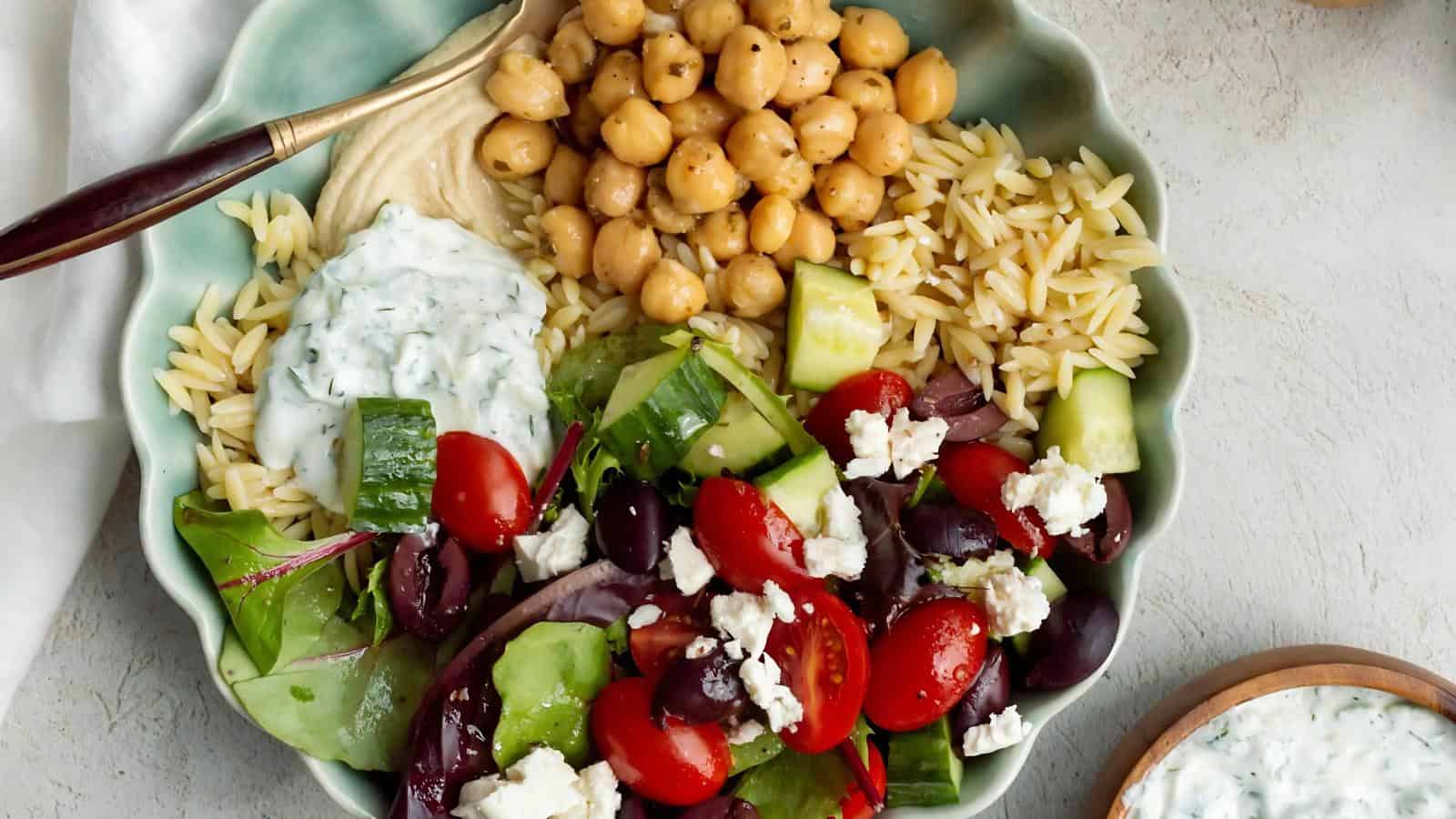 A plate with orzo, chickpeas, hummus, cucumbers, cherry tomatoes, olives, feta cheese, and mixed greens, topped with tzatziki sauce, served with a fork.