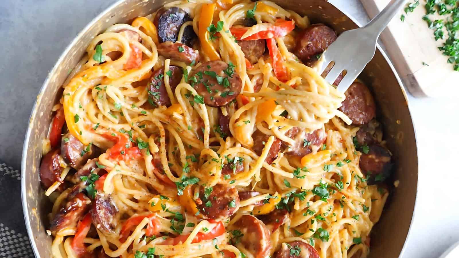 A skillet filled with spaghetti, sliced sausage, and colorful bell peppers, garnished with chopped parsley.