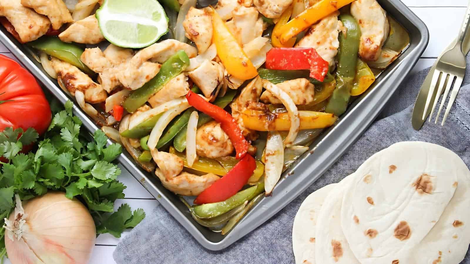 A tray of grilled chicken and mixed bell peppers next to lime slices. Fresh cilantro, a tomato, an onion, and tortillas are on the side. White surface underneath.