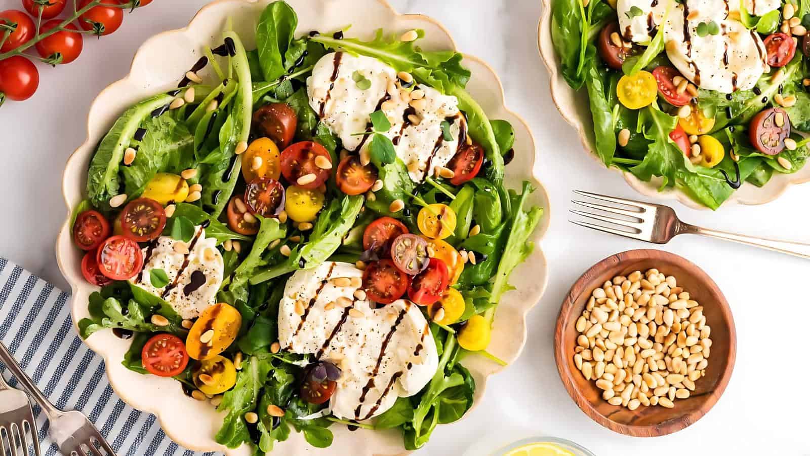 A salad with tomatoes, mozzarella, and mixed greens drizzled with balsamic glaze, accompanied by a small bowl of pine nuts.