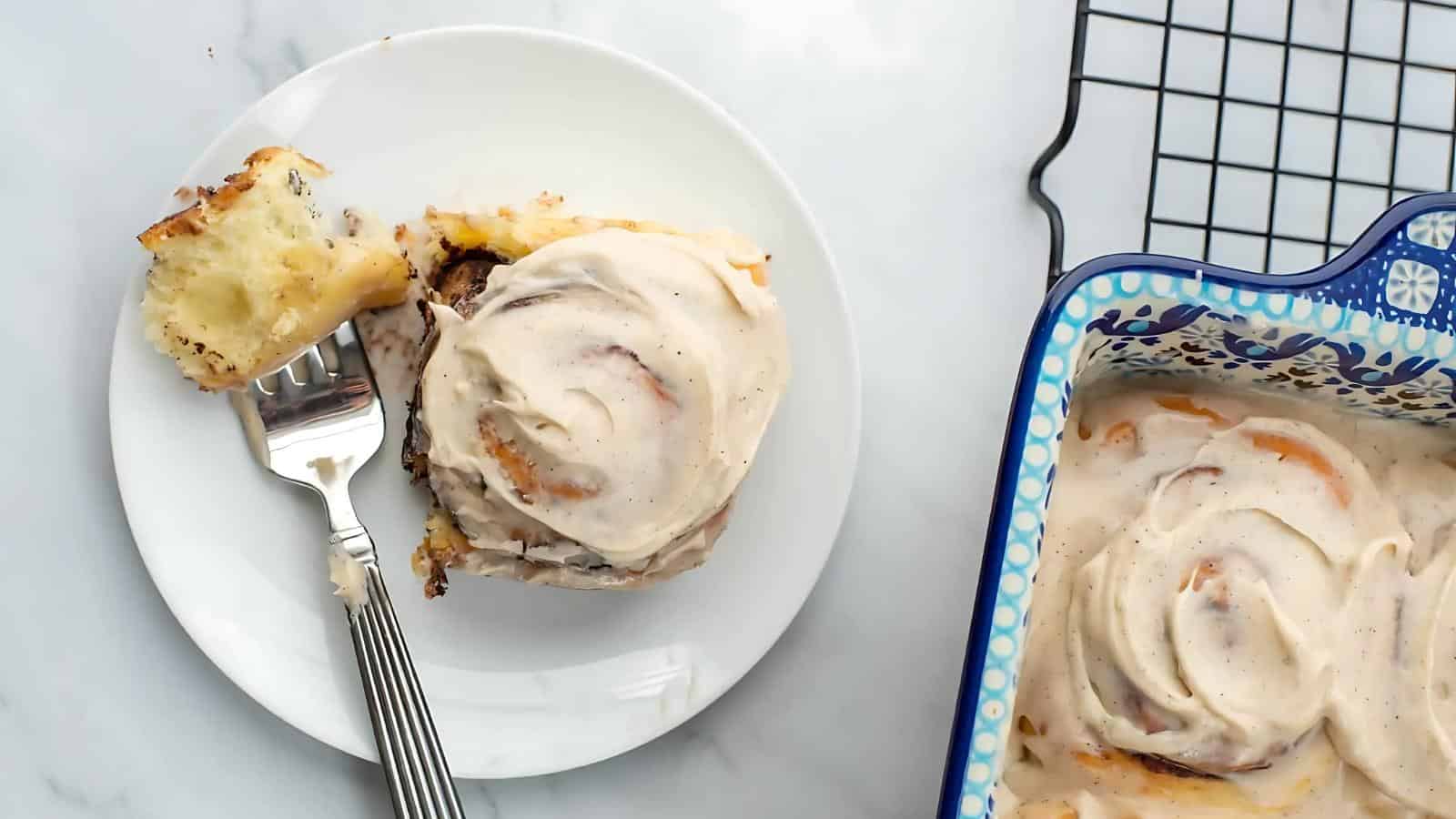 Cinnamon roll with icing on a white plate, partially eaten with a fork. A ceramic dish with more cinnamon rolls is nearby on a cooling rack.