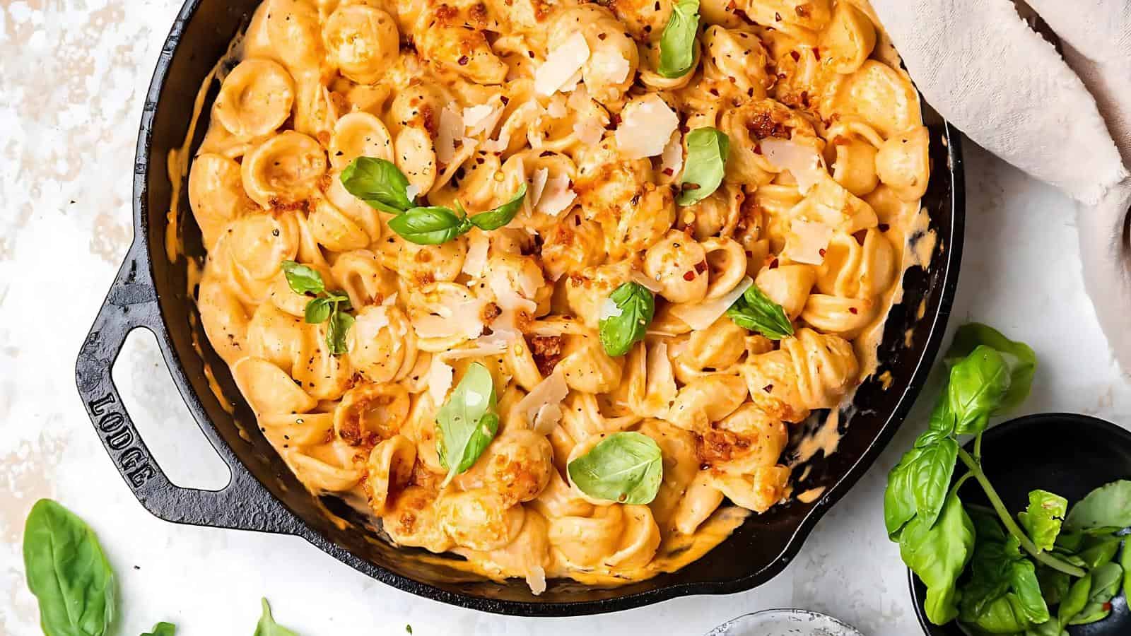 A skillet filled with creamy pasta garnished with fresh basil leaves and grated cheese, placed on a light-colored surface with a few sprigs of basil nearby.