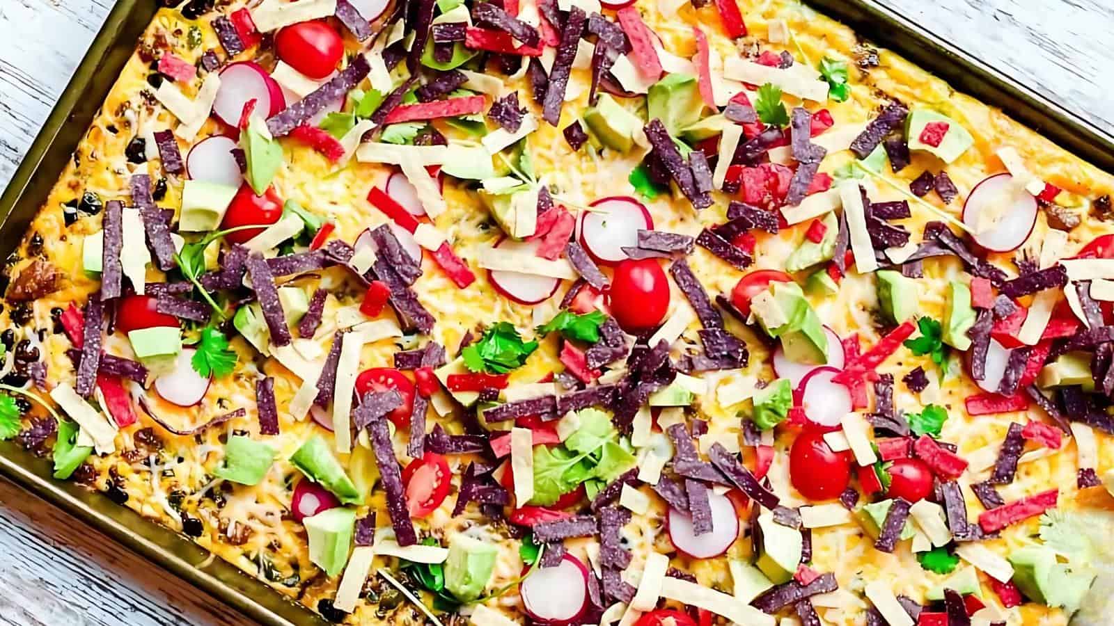 A colorful taco salad topped with cherry tomatoes, radish slices, avocado chunks, shredded cheese, and crispy tortilla strips on a rectangular tray.